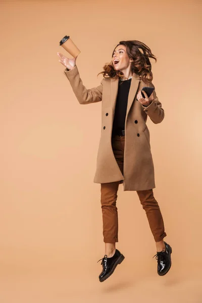 优雅的女人20多岁的图像穿着外套举行手机和外卖咖啡 而走隔离在米色背景 — 图库照片