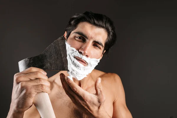 Image of handsome young naked man posing isolated over dark wall background with shaving gel on beard shaving his beard with axe.