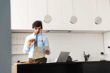 Yakışıklı genç iş adamı mutfak at resmini gazetede kulaklıklar giyen dizüstü bilgisayar kullanan sandviç yemek.