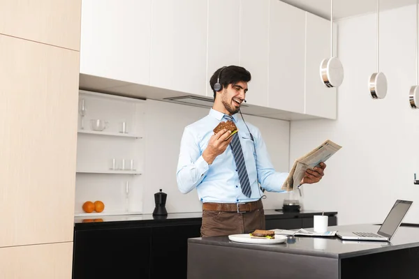 Image of a happy laughing young business man at the kitchen eat sandwich using laptop computer wearing headphones reading newspaper.