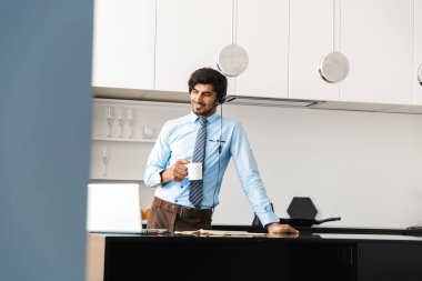 Evde mutfakta çalışan, dizüstü bilgisayar kullanan, kulaklık kümesi aracılığıyla iletişim, kahve içme kendine güvenen genç işadamı