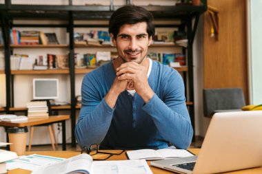 Çalışma/eğitim, kullanarak dizüstü bilgisayar kitaplığı masada oturan emin yakışıklı genç
