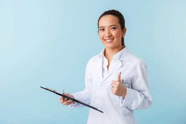 陽気な若い女性医師親指を示す青い壁背景持株クリップボード上孤立したポーズの写真 — ストック写真