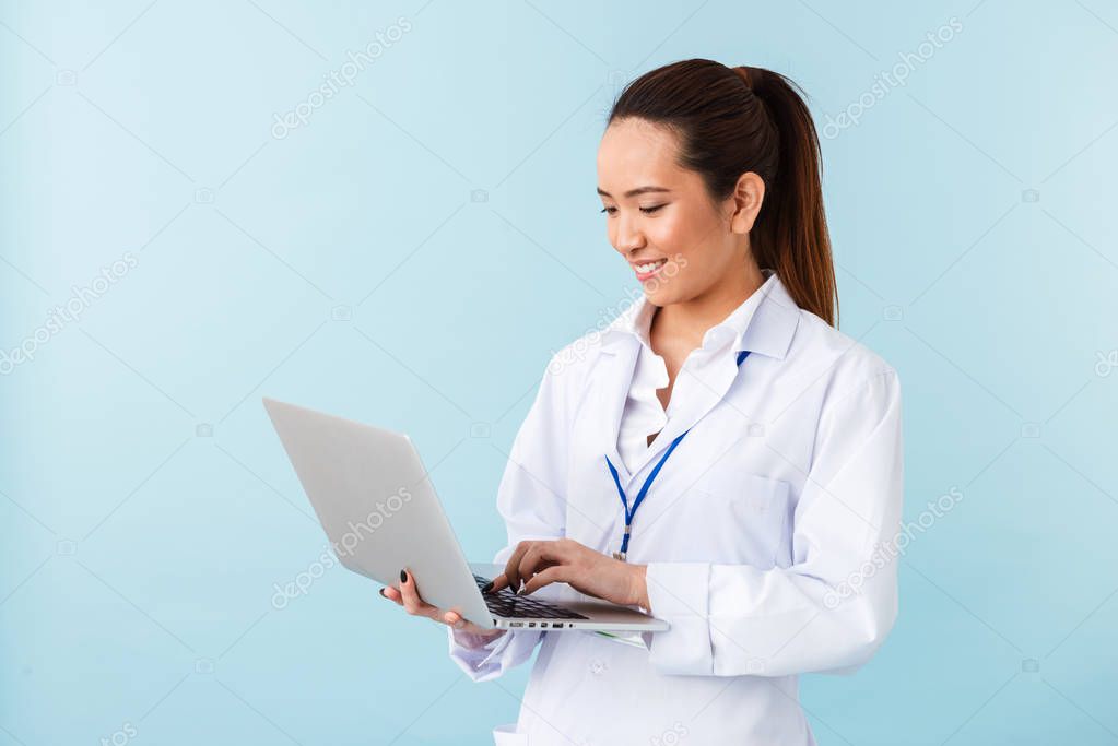 Photo of a young woman doctor posing isolated over blue wall background using laptop computer.