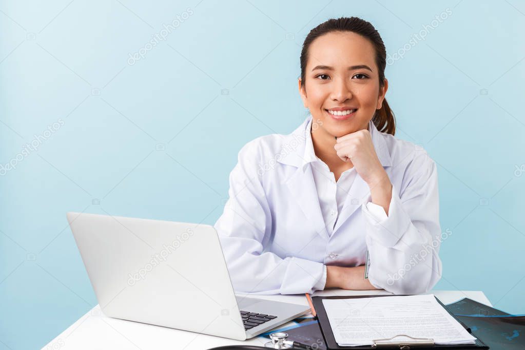 Photo of a young woman doctor posing isolated over blue wall background using laptop computer.