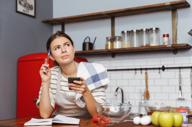 Mutlu genç kadın evde mutfakta ayakta, yemek tarifi kitabı ve cep telefonu ile kahvaltı