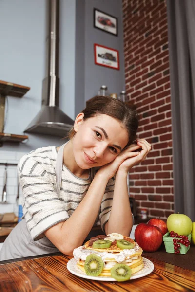 快乐的年轻女子在厨房在家里 显示准备好的煎饼与水果和奶油在桌子上 — 图库照片
