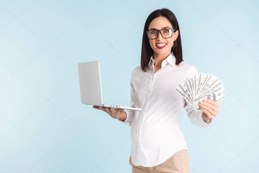 Image of a beautiful young pregnant business woman isolated over blue wall background using laptop computer holding money.