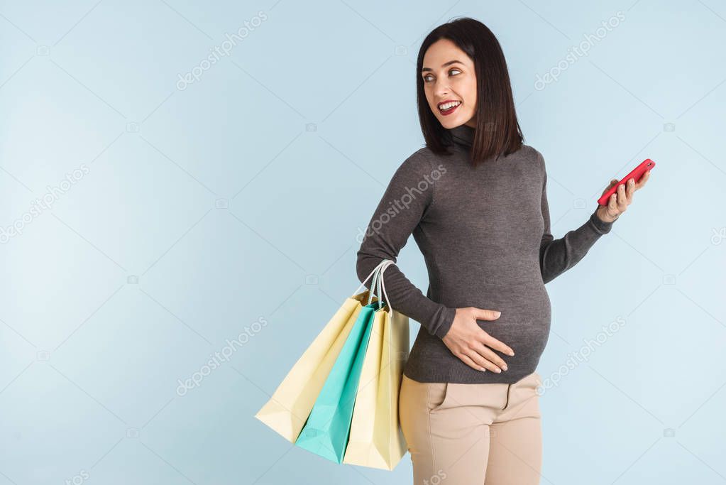 Photo of a young pregnant woman isolated over blue wall background using mobile phone holding shopping bags.