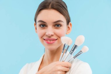 Makyaj fırçaları tutan mavi arka plan üzerinde izole poz güzel genç kadın fotoğrafı.