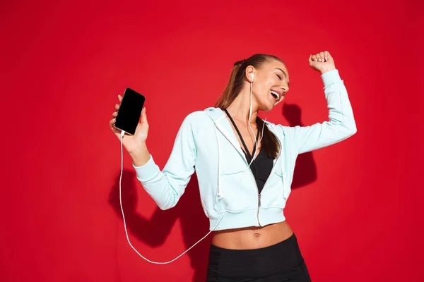 一个美丽健康的年轻女运动员站在红色背景 听音乐与耳机 拿着手机的肖像 — 图库照片