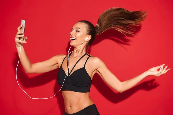 一个美丽健康的年轻女运动员的肖像跳过红色的背景 听音乐用耳机 拿着手机 — 图库照片