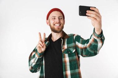 Yakışıklı adam şapka ve beyaz arka plan üzerinde izole ayakta iken cep telefonundan selfie fotoğraf çekme ekose gömlek giymiş görüntüsünü