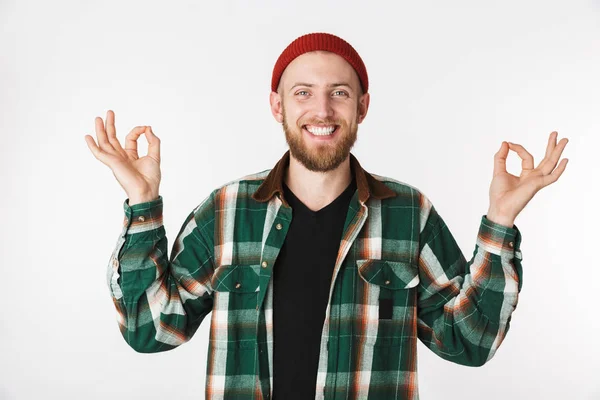 Portrait of smiling bearded guy wearing hat and plaid shirt showing meditation sign while standing isolated over white background