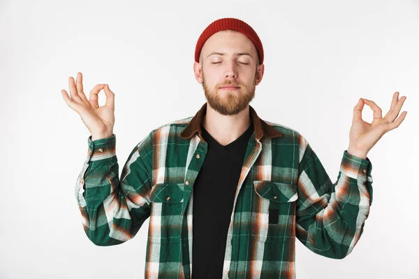 Portrait of joyous bearded guy wearing hat and plaid shirt showing meditation sign while standing isolated over white background