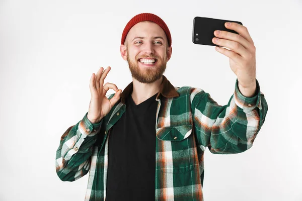 高兴的家伙戴着帽子和格子衬衫在手机上自拍照片的图像 而站在孤立的白色背景 — 图库照片