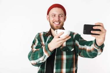 Fincan çay ile tutarak ve beyaz arka plan üzerinde izole ayakta iken cep telefonunuzu kullanarak kareli gömlek giyen olumlu adam portresi