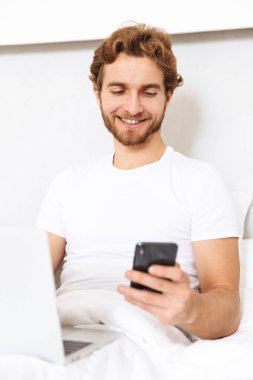 Yakışıklı bir genç sakallı adam yatakta görüntüsünü evde dizüstü bilgisayar ve cep telefonu kullanma.
