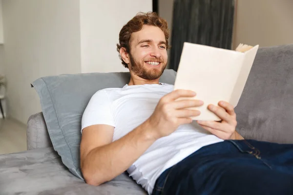 自宅のソファーにハンサムな若いひげを生やした男のイメージは 本を読んでください — ストック写真