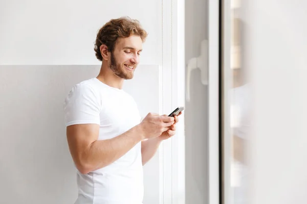 Leende Ung Man Håller Mobiltelefon Står Vid Fönstret Hemma — Stockfoto