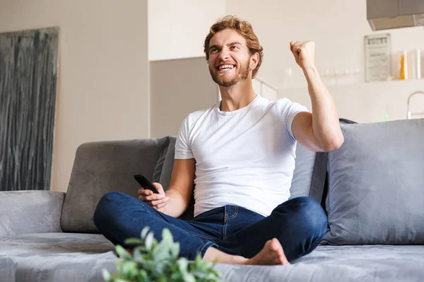 Foto Guapo Joven Emocional Sofá Casa Sosteniendo Control Remoto Ver —  Fotos de Stock