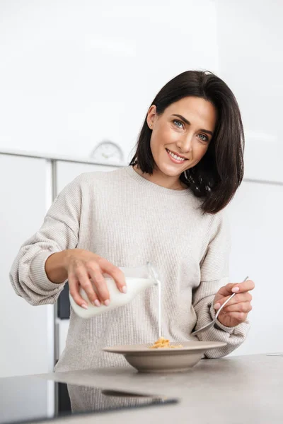 Image of kind woman 30s making breakfast with oatmeal and fruits while standing in modern kitchen at home