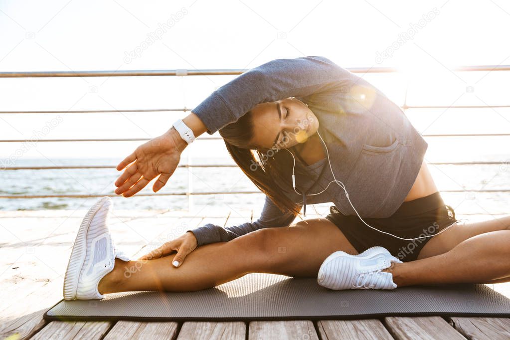 Image of a beautiful young sports fitness woman make stretching exercises at the beach outdoors listening music with earphones.