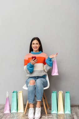 Görüntü güzel kadının 30'lu çanta alışveriş ve cep telefonuyla renkli sandalye ile oturan gri arka plan üzerinde izole