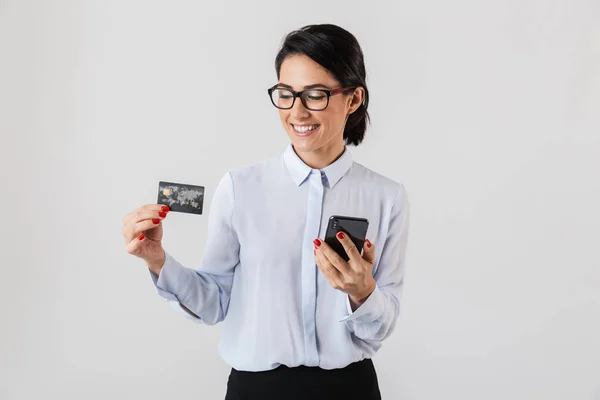 携帯電話と白い背景上分離されたクレジット カードを保持している眼鏡を着て自信を持って事務所女性のイメージ — ストック写真