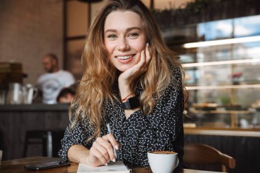 Güzel genç güzel kadın kapalı kahve içme not defterinde notlar yazma kafede oturan fotoğrafı. Görünümlü kamera.