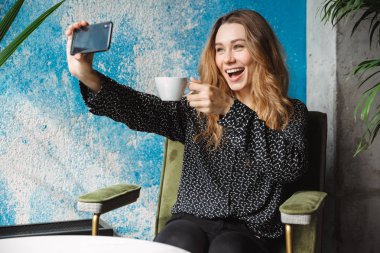 Kahve içme cep telefonuyla bir selfie almak kapalı kafede oturan mutlu duygusal güzel genç güzel kadın fotoğraf.