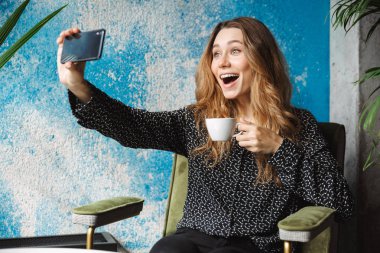 Kahve içme cep telefonuyla bir selfie almak kapalı kafede oturan mutlu duygusal güzel genç güzel kadın fotoğraf.