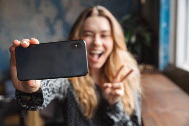 Kapalı kafede oturan şok heyecanlı güzel genç güzel kadın fotoğrafını çekin selfie cep telefonuyla.