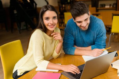 Genç öğrenciler arkadaş çift okumak ev ödevi eğitim ve dizüstü bilgisayar kullanan Kitaplığı'nda.