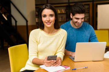 Genç öğrenciler arkadaş çift okumak ev ödevi eğitim ve dizüstü bilgisayarı ve cep telefonu kullanarak Kitaplığı'nda.