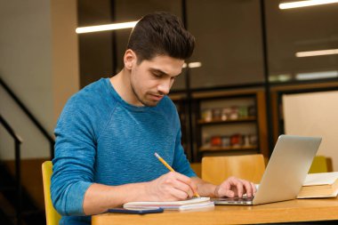 Genç bir öğrencinin görüntü okuma eğitimi ve dizüstü bilgisayar kullanan ödev Kütüphane erkekte konsantre.