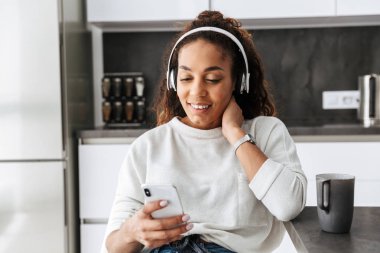 Parlak mutfakta otururken smartphone kullanarak kulaklık giyen mutlu Afro-Amerikan kız resim