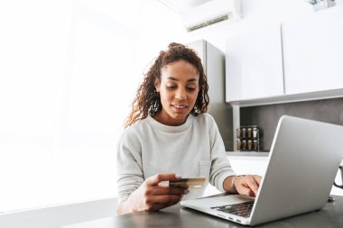 Aydınlık oturma odası masada otururken dizüstü bilgisayar ve kredi kartı kullanan memnun Afro-Amerikan kız resim