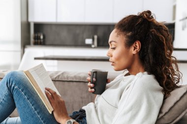 Kitap okuma ve parlak düz kanepede otururken çay içme sevimli Afro-Amerikan kız resim