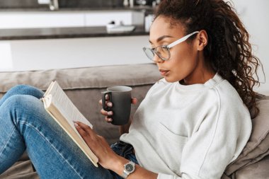 Kitap okuma ve parlak düz kanepede otururken çay içme akıllı Afro-Amerikan kız resim