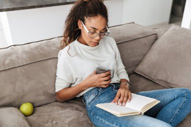 Kitap okuma ve parlak düz kanepede otururken çay içme çekici Afro-Amerikan kız resim