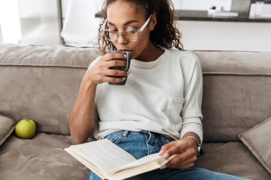 Kitap okuma ve parlak düz kanepede otururken çay içme esmer Afro-Amerikan kız resim