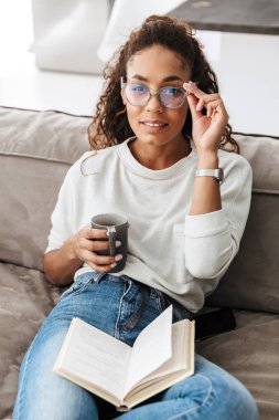 Kitap okuma ve parlak düz kanepede otururken çay içme şık Afro-Amerikan kız resim