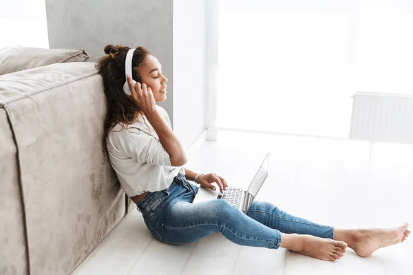 明るい居間の床に座ってラップトップを使用してヘッドフォンを身に着けている美しいアフリカ系アメリカ人女性の写真 — ストック写真