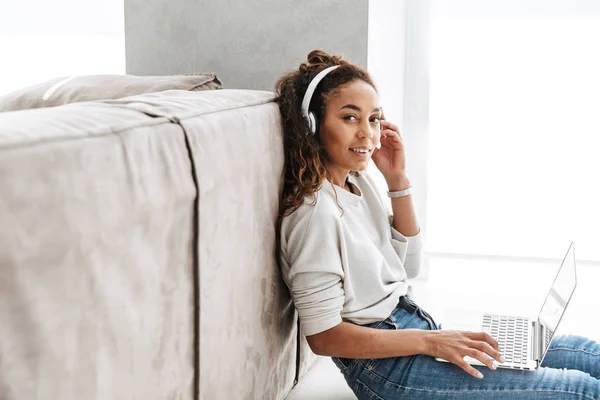 明るい居間の床に座ってラップトップを使用してヘッドフォンを身に着けている現代のアフリカ系アメリカ人女性の写真 — ストック写真