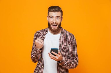 Cep telefonu kullaranarak sarı arka plan üzerinde izole ayakta rahat kıyafetler giymiş neşeli genç bir adam portresi