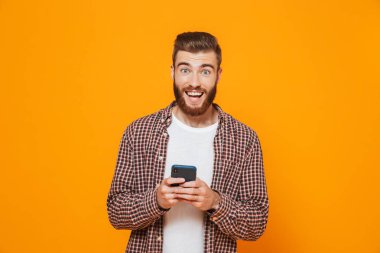 Cep telefonu kullaranarak sarı arka plan üzerinde izole ayakta rahat kıyafetler giymiş neşeli genç bir adam portresi