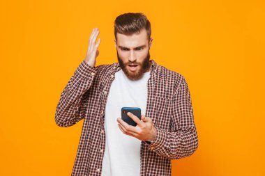 Cep telefonu kullaranarak sarı arka plan üzerinde izole ayakta rahat kıyafetler giymiş karışık genç bir adam portresi