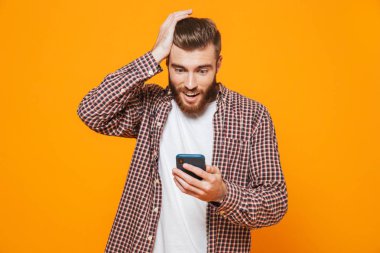 Cep telefonu kullaranarak sarı arka plan üzerinde izole ayakta rahat kıyafetler giymiş karışık genç bir adam portresi
