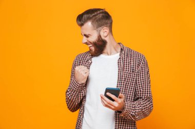 Cep telefonu kullaranarak sarı arka plan üzerinde izole ayakta rahat kıyafetler giymiş neşeli genç bir adam portresi